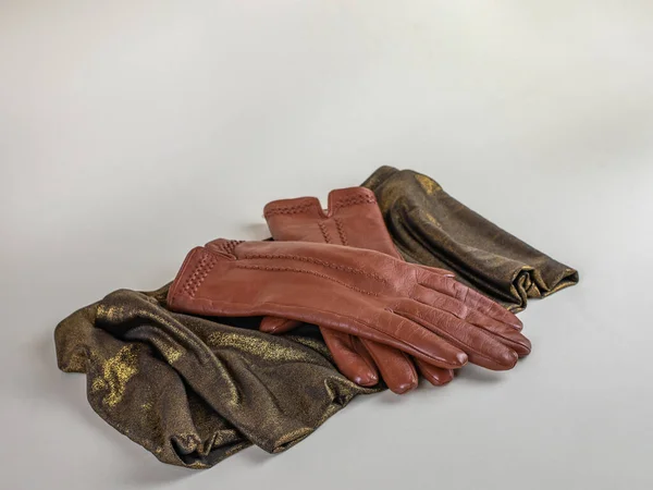 A pair of new brown leather gloves on a green and gold scarf, isolated on a light background. Graceful womens brown leather gloves lying on a green scarf with a golden sheen on a light background.