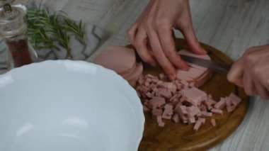 Boiled sausage is cut into small pieces with a kitchen knife on a wooden cutting board for making meat salad. Pepper grinder, sprig of rosemary next to the cutting board. Womens hands close up