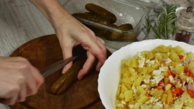 Housewife cuts pickled cucumbers into small cubes with knife on wooden cutting board on gray kitchen table. Close-up female hand cuts pickled cucumbers for vegetable salad. Concept of vigaterianism.