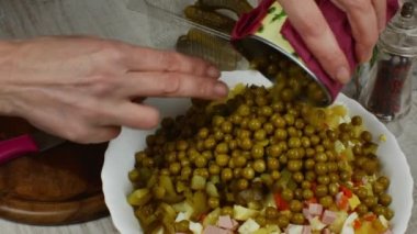 Canned green peas are sprinkled into a bowl of chopped vegetables to prepare a meat salad, next to it on a gray kitchen table is a wooden cutting board, a pepper grinder, a sprig of rosemary.