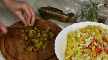 Housewife cuts green pickled cucumbers with a kitchen knife on a wooden cutting board and puts the cut cucumbers in a bowl for preparing a vegetable salad. Female hands close up. Concept of veganism.