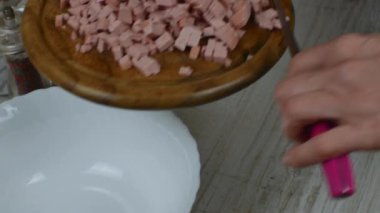 Tahta kesme tahtasından küçük haşlanmış sosis parçaları bıçakla beyaz cam kaseye dökülüyor. Kadınların elleri ahşap kesme tahtasından yapılmış bıçakla haşlanmış sosis döker..