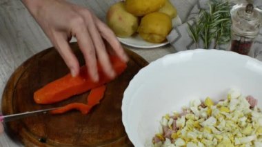 Housewife cuts boiled carrots with knife on wooden cutting board on kitchen table, next to it are bowl of meat salad being prepared, saucer with boiled potatoes, pepper grinder, sprig of rosemary.