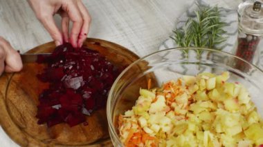 Housewife cuts red boiled beets with a kitchen knife on a wooden cutting board and puts the cut cubes of beets in a bowl for preparing vegetable salad. Female hands close up. The concept of veganism.