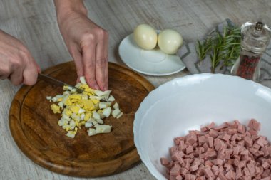 A housewife cuts a boiled egg finely on a wooden cutting board for making meat salad. Close-up of female hands slicing a boiled egg on a wooden cutting board with a kitchen knife.
