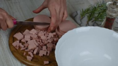 Sosis bıçakla tahtayı keserken küçük parçalara ayrılır. Kadın elleri, gri mutfak masasında ahşap kesme tahtasının üzerinde bıçakla haşlanmış sosisi ince ince doğruyor. Et salatası hazırlığı.