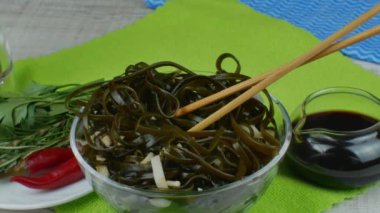 Seaweed kelp salad with onion and soy sauce in a bowl and wooden chopsticks. On a green napkin there is a bowl of seaweed salad, saucers with red hot peppers, herbs and a gravy bowl with soy sauce.