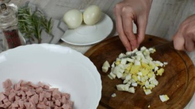 Haşlanmış tavuk yumurtası ahşap kesim tahtasıyla mutfak bıçağıyla kesilir ve doğranmış yumurta, doğranmış sosisle birlikte cam bir kaseye yerleştirilir. Et salatası hazırlayan bir ev kadınının yakın çekimi..