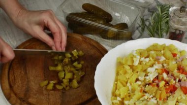 Pickled cucumbers are cut into small pieces with kitchen knife on cutting board, next to it are bowl of vegetable salad being prepared, box of pickled cucumbers, pepper grinder, a sprig of rosemary