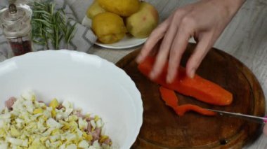 Kaynamış havuçlar ahşap bir kesim tahtasının üzerinde bıçakla kesilir, yanında et salatası, haşlanmış patatesli çay tabağı, biber öğütücü, biberiye filizi bulunur. Kadın elleri yakın..