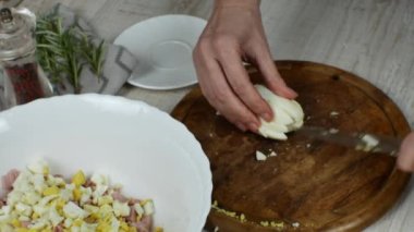 A housewife cuts a boiled chicken egg finely on a wooden cutting board and pushes the egg with a knife into a white glass bowl for making meat salad. Close-up of female hands preparing meat salad.