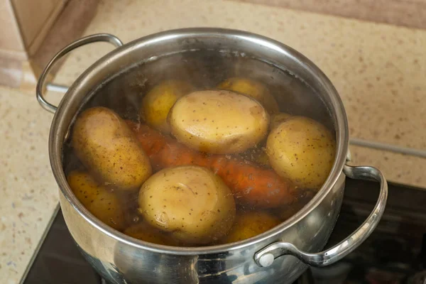 Metal pan without a stainless steel lid on a home gas stove boils potatoes and carrots in boiling water with bubbles. The process of cooking boiled potatoes and carrots in the home kitchen. Close-up.