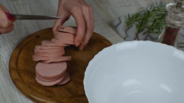 Dilimlenmiş sosis dilimleri ahşap bir kesme tahtasına konur ve et salatası hazırlamak için küçük parçalara ayrılır. Biber öğütücü, biberiye filizi kesme tahtasının yanında. Kadınların elleri kapalı..