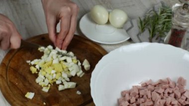 A housewife cuts a boiled chicken egg finely on a wooden cutting board and pushes the egg with a knife into a white glass bowl for making meat salad. Close-up of female hands preparing meat salad.
