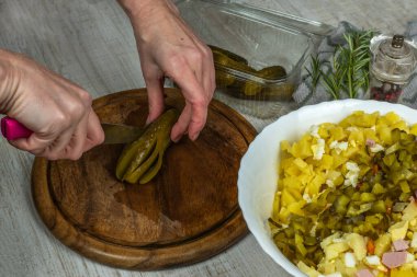 Housewife cuts pickled cucumbers into small cubes with knife on wooden cutting board on gray kitchen table. Close-up female hand cuts pickled cucumbers for vegetable salad. Concept of vigaterianism.