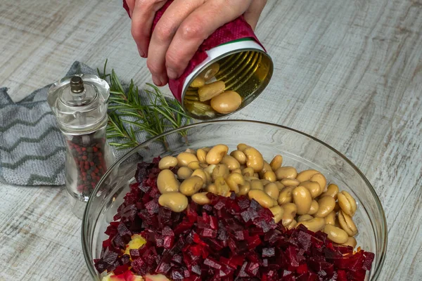 Housewife sprinkles canned white beans from iron can into a bowl on gray kitchen table. Close-up female hand sprinkles white canned beans into bowl for preparing vegetable salad. Concept of veganism.