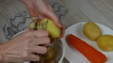 Removing skin from boiled potatoes. Housewife peels yellow boiled potatoes with iron table knife and puts the peeled potatoes in white glass bowl. Vegetarian food concept. Close-up from the top side.