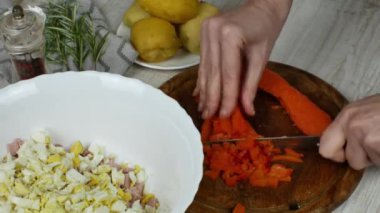 Housewife cuts boiled carrots with knife on wooden cutting board on kitchen table, next to it are bowl of meat salad being prepared, saucer with boiled potatoes, pepper grinder, sprig of rosemary