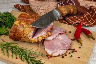 Traditional ham sliced on a wooden cutting board with a carving knife, multi-colored allspice, red hot peppers, parsley, dill and basil. The concept of snacks from pork meat.