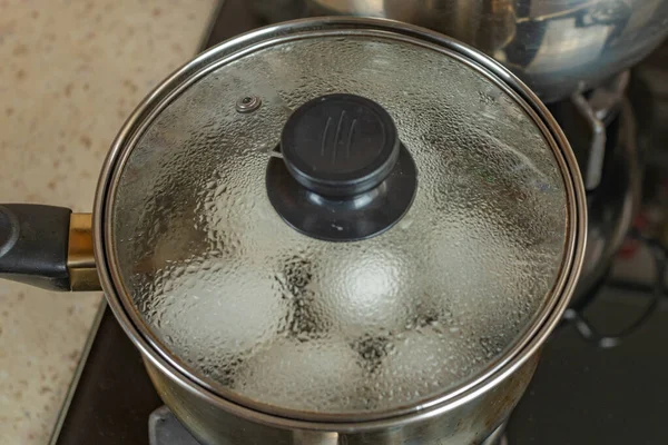 stock image White chicken eggs are boiled in a stainless steel iron saucepan with a transparent glass lid. White chicken eggs are boiled in bubbling hot water in a saucepan on a gas stove. Close-up top view.