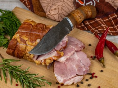 Traditional ham sliced on a wooden cutting board with a carving knife, multi-colored allspice, red hot peppers, parsley, dill and basil. The concept of snacks from pork meat.