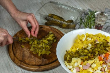 Housewife cuts pickled cucumbers into small cubes with knife on wooden cutting board on gray kitchen table. Close-up female hand cuts pickled cucumbers for vegetable salad. Concept of vigaterianism.