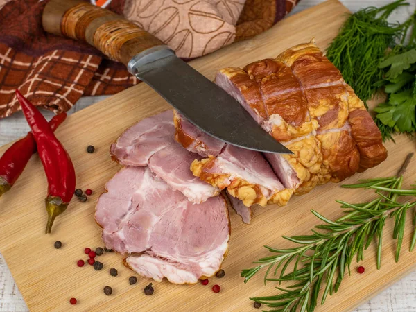 Traditional ham sliced on a wooden cutting board with a carving knife, multi-colored allspice, red hot peppers, parsley, dill and basil. The concept of snacks from pork meat.