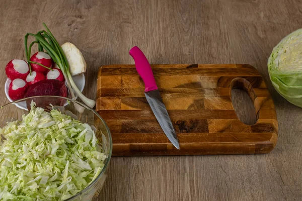 Salata için sebze seti mutfak masasındaki tahta kesme tahtasının üzerinde yatıyor. Beyaz lahana, kırmızı turp, yeşil soğan, kırmızı pancar kökü, kereviz kökü, kesme tahtası, bıçak ve cam kase mutfak masasında..