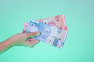 Hand holding replica Indonesian currency, 100,000 and 50,000 rupiah banknotes on cyan background