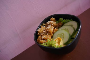 Lezzetli tavuklu teriyaki kasesi susam tohumları, marul ve salatalık dilimleri kahverengi bir masada.