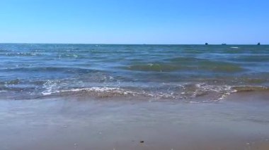 Sakin deniz dalgalarının berrak mavi bir gökyüzünün altında kumlu bir plaja yuvarlanışının huzurlu bir görüntüsü. Arkaplan, rahatlama veya kıyı temaları için ideal.