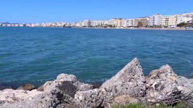 Mavi sulara, kayalık ön plana ve berrak bir gökyüzünün altındaki uzak sıradağlara uzanan ahşap bir iskeleyi gösteren sakin bir sahne..