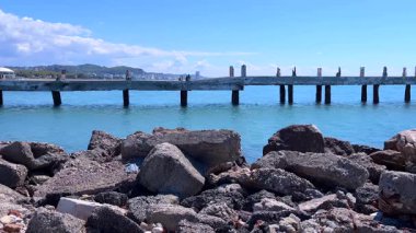 Sakin deniz dalgalarının ön planda kayalık oluşumlara çarptığı geniş bir kıyı manzarası ve parlak mavi gökyüzünün altında uzak bir şehir silüeti..