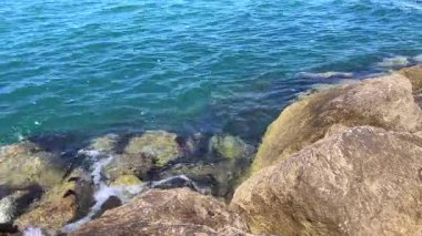 Parlak ve sakin deniz görüntüleri kayalık oluşumlara hafifçe sürtünüyor, batık taşları ve güneş yansımalarını ortaya çıkarıyor..