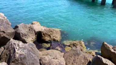 Temiz turkuaz deniz dalgaları eski bir ahşap iskelenin altındaki kayalık sahile nazikçe çarpıyor. Su yansımaları ve kırsal dokularla huzurlu bir sahil manzarası..