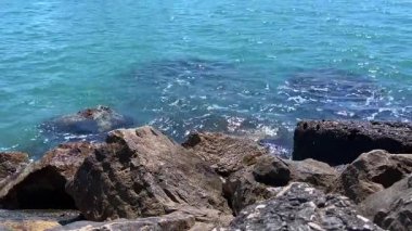 Parlak bir turkuaz deniz, karanlık, desenli kıyı şeridi taşlarına hafifçe çarpar ve ışık ve doğal yapının zıtlığını yaratır..