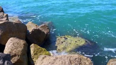 Mavi deniz suyunun sıcak tonlu kayalara hafifçe sürtündüğü yakın çekim videosu doğal dokuları ve hareketleri vurguluyor..