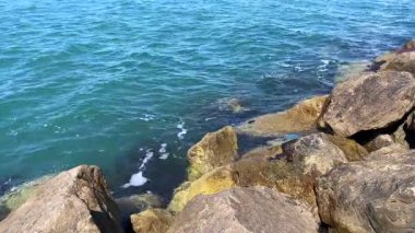 Güneşli bir günde mavi okyanus dalgalarının büyük kıyı kayaları arasında yavaşça dolaştığı sakin bir sahil sahnesi..