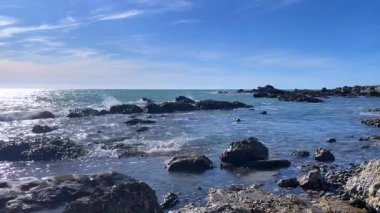 Sakin bir okyanusun sakin bir görüntüsü. Yukarıdaki açık mavi gökyüzü ve barışçıl dalgalarla kayalık bir kıyıya hafifçe çarpıyor..