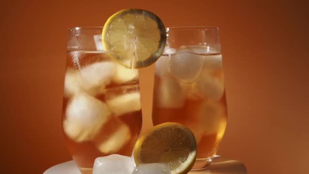 アイスティーをオレンジの背景に氷のキューブでガラスに回転させます 冷たいお茶と氷 — ストック動画