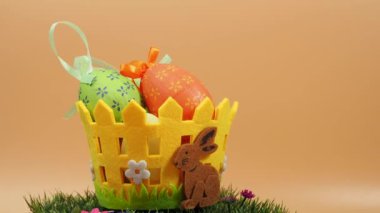 Yeşil çimlerin üzerinde dekoratif yumurtalar bulunan Paskalya sepeti turuncu arka planda döner. Paskalya dekorasyonu.