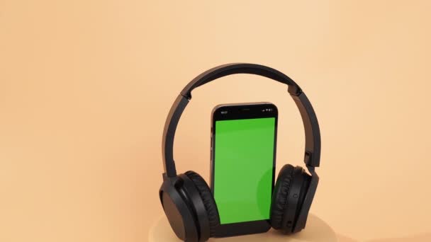 黑色无线耳机和带有绿色屏幕的智能手机在橙色背景下旋转 无线耳机特写 — 图库视频影像