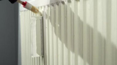 Bir işçi fırça kullanarak ısıtma kaloriferine bir kat boya sürer. Isıtma ünitesini boyuyorum..
