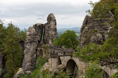 Bir grup insan kayalık bir bölgenin üzerindeki köprüden geçiyor. Köprü taştan yapılmıştır ve ağaçlarla çevrilidir.