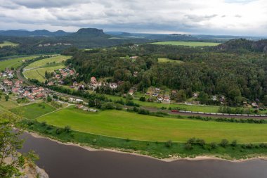 Bir kasaba ve tren geçen bir nehir manzarası. Şehir yeşil bir tarla ile çevrilidir ve nehir kahverengidir.