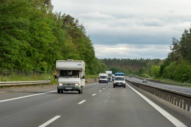 Almanya, 17 Mayıs 2024. Beyaz bir karavan diğer araçlarla birlikte otoyolda ilerliyor. Sahne sakin ve huzurlu, ağaçlar yol boyunca ve gökyüzü yukarıda..