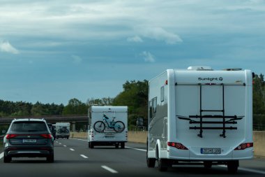 Almanya, 17 Mayıs 2024. Üstünde bisiklet olan beyaz bir karavan yoldan aşağı gidiyor. Römork bir araba ve başka bir karavanın yanında..