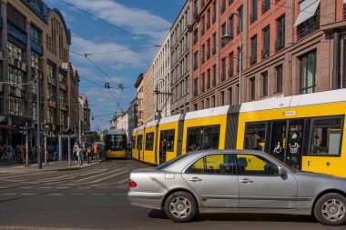 Almanya 17 Mayıs 2024. Gümüş renkli bir araba sarı bir trenin arka planına doğru gidiyor. Hayat tam gaz devam ediyor: insanlar yürüyor ve bisiklete biniyor. Sahne canlı ve enerjik.
