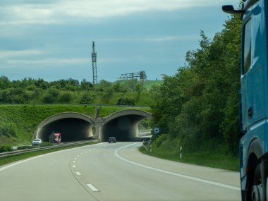 17 Mayıs 2024 'te bir mavi römork arka planda bir tünelle yol boyunca ilerliyor. Tünel ağaçlarla çevrili ve gökyüzü bulutlu..