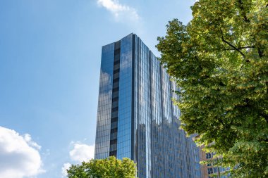 Bir sürü penceresi ve arka planda bulutlu bir gökyüzü olan yüksek bir bina. Binanın önündeki ağaçlar yeşil ve cam yapıya güzel bir kontrastlık sağlıyor.